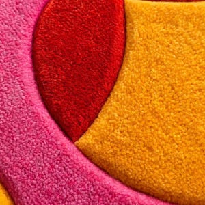 May include: A tufted textile rug featuring a geometric pattern with distinct sections of vibrant colors. The design includes a large curved segment in bright red, a textured area in deep pink, and a prominent section in golden yellow.
