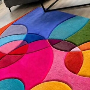 May include: A plush area rug featuring a vibrant, multi-colored geometric pattern composed of various circular and curved shapes. The design includes bold shades of hot pink, bright orange, deep red, sunny yellow, sky blue, and forest green.