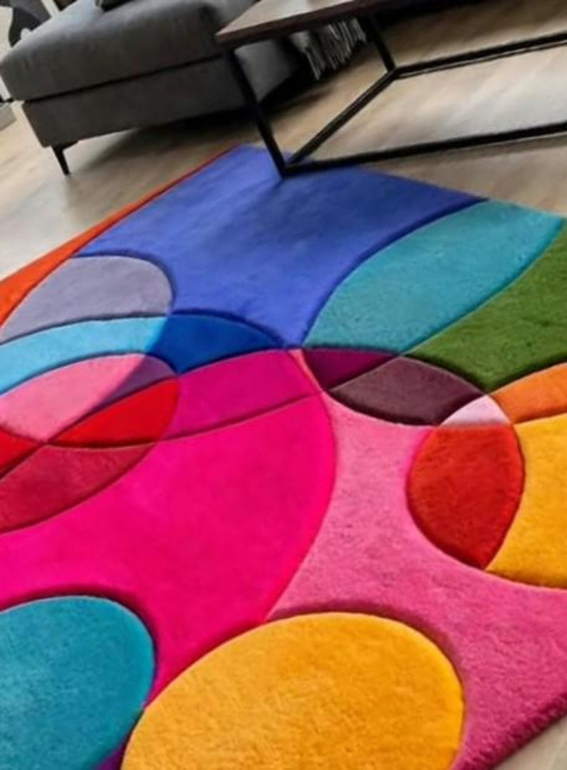 May include: A plush area rug featuring a vibrant, multi-colored geometric pattern composed of various circular and curved shapes. The design includes bold shades of hot pink, bright orange, deep red, sunny yellow, sky blue, and forest green.