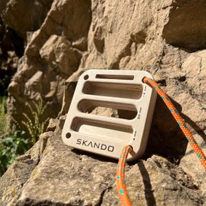 May include: A square light-colored wooden climbing hangboard with three horizontal slots and rounded corners. The board features the text SKANDO and labels for 4 mm, 20 mm, and 12 mm depths. An orange and teal climbing rope is threaded through the holes.