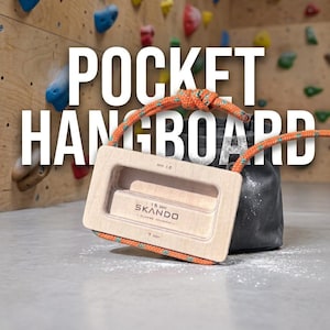 Op de afbeelding: Een lichtgekleurd houten pocket hangboard met een bevestigd oranje klimtouw. Het bord bevat gegraveerde tekst met 21 mm, 15 mm, 7 mm en SKANDO CLIMBING EQUIPMENT. Een donkergrijze pofzak staat direct achter het hangboard.