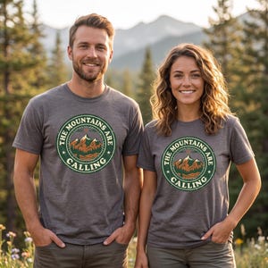Puede incluir: Una camiseta gris carbón de manga corta con un gráfico circular en el pecho. El diseño incluye una ilustración de una cordillera, un bosque y el texto "THE MOUNTAINS ARE CALLING" dentro de un borde verde de estilo vintage.