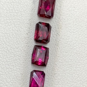 May include: Four rectangular faceted gemstones with a deep magenta and red hue arranged in a vertical line. Each stone features a clear, polished surface with multiple geometric facets that reflect light, showcasing a vibrant, saturated color.