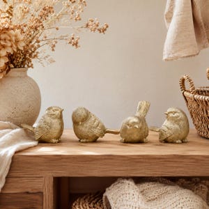 Puede incluir: Cuatro pequeñas figuras de pájaros de color dorado están dispuestas en fila. Cada pájaro presenta una superficie texturizada y tallas detalladas en las alas. Las figuras están colocadas sobre una superficie de madera con acabado metálico.