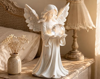 May include: A white angel figurine with detailed feathered wings and flowing robes, holding a glowing, clear spherical orb in its hands. The sculpture features a serene facial expression and stands upright with a smooth, matte finish.
