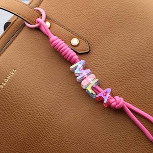 Puede incluir: Un llavero de cordón rosa brillante con cuentas de letras iridiscentes y translúcidas que forman el nombre MILA. El cordón está anudado firmemente y sujeto a un bolso de cuero color canela con herrajes dorados y el texto POPPY & PEONIES.