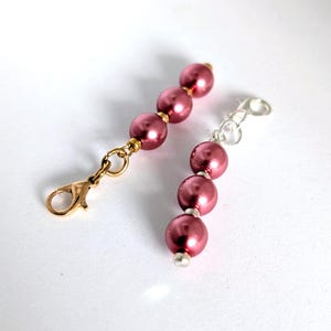May include: Two beaded zip pulls featuring round, dark pink faux pearls. One pull has gold-toned metal hardware with a lobster claw clasp, while the other pull features silver-toned metal hardware with a matching lobster claw clasp attachment.