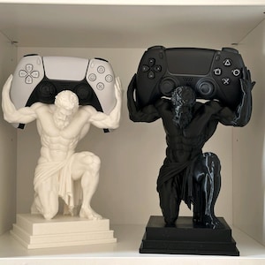 Könnte beinhalten: Zwei Statuen einer knienden muskulösen Figur, eine weiß und eine schwarz, die jeweils einen Gamecontroller über dem Kopf halten. Die weiße Statue trägt einen weiß-schwarzen Controller, während die schwarze Statue einen schwarzen Controller hält.