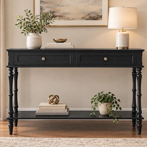 May include: A black rectangular console table featuring two drawers with small round brass-toned knobs. The table is supported by four turned legs and includes a lower shelf for storage. The surface is smooth and finished in a matte black paint.