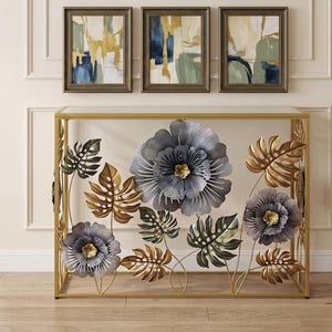 May include: A rectangular console table with a gold metal frame and a glass top. The front panel features decorative metal floral and monstera leaf motifs in shades of metallic gold, dark gray, and silver, with three framed abstract art prints above.