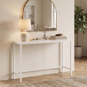 May include: A white rectangular console table with a slim metal frame. On the table sits a beige lamp with a cylindrical shade, a rectangular tray with three smooth stones, a glass vase with dried branches, and a stack of books with a bowl on top.