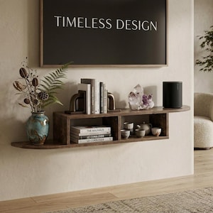 May include: A dark wood floating wall shelf featuring two rectangular compartments. The shelf holds a blue ceramic vase with dried foliage, books, bookends, a large amethyst crystal cluster, and a black cylindrical speaker. Text reads TIMELESS DESIGN.