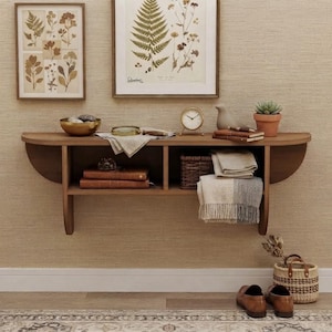 May include: A wall-mounted wooden shelf with two compartments holding books, a woven basket, and folded textiles. On top sits a gold-rimmed bowl, a magnifying glass, a small clock, a bird figurine, and stacked notebooks. A woven basket and shoes sit below.