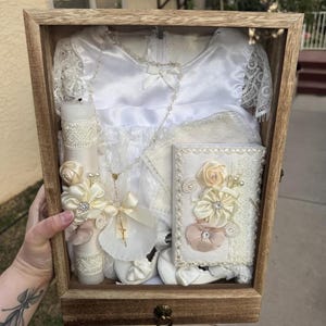 May include: A wooden display box containing a white christening gown with lace sleeves, a matching candle adorned with cream fabric flowers, a small white book decorated with floral accents, a pair of white shoes, and a delicate beaded rosary.