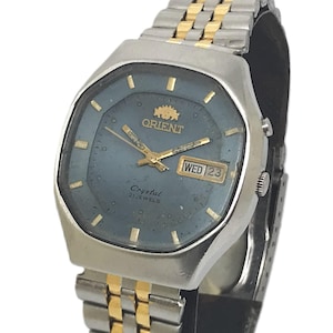 May include: An Orient wristwatch with a blue octagonal dial, gold-tone hands, and hour markers. The watch features a day-date window at the three o'clock position, the text Orient Crystal 21 Jewels on the face, and a two-tone silver and gold metal link band.