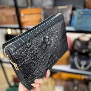 Op de afbeelding: Een zwarte rechthoekige clutch met een gestructureerd krokodillenleerpatroon. De tas heeft een goudkleurige ritssluiting aan de bovenkant en een goudkleurige metalen sluiting aan de zijkant. De tekst Lucky Strike Handmade is onderaan zichtbaar.