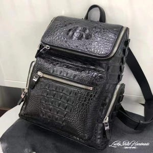 May include: A black backpack featuring a textured crocodile skin pattern. The bag includes a top flap, a front zippered pocket, and side zippered compartments, all accented with silver-toned metal zippers. The text Lucky Strike Handmade is visible.