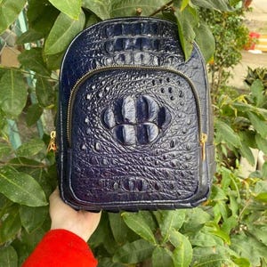 Puede incluir: Una mochila de color azul oscuro con un patrón texturizado de piel de cocodrilo. La bolsa tiene una parte superior redondeada, un bolsillo frontal con cremallera dorada y un panel central con escamas más grandes y prominentes.