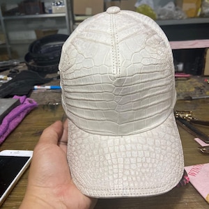May include: A white baseball cap featuring a textured, embossed pattern that mimics the appearance of crocodile skin. The cap has a structured crown and a curved brim, with visible stitching along the panels and the edge of the visor.