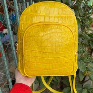 Puede incluir: Una mochila de color amarillo brillante con un patrón texturizado de piel de cocodrilo. La bolsa tiene una parte superior redondeada, un bolsillo frontal grande con cremallera y correas amarillas a juego. El texto Lucky Strike Handmade es visible en la esquina inferior derecha.