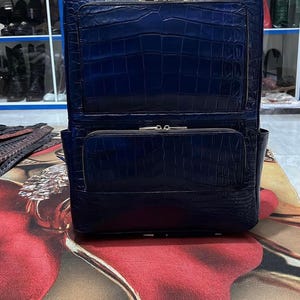 Puede incluir: Una mochila de color azul profundo con un patrón de textura de piel de cocodrilo. La bolsa incluye un asa superior, un compartimento principal grande con cierre de cremallera y un bolsillo frontal más pequeño con cremallera y herrajes.