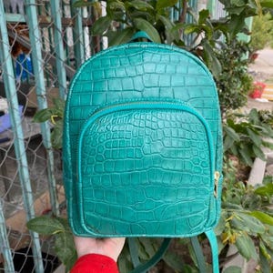 Puede incluir: Una mochila de color verde azulado con un patrón texturizado de piel de cocodrilo. La bolsa tiene una parte superior redondeada, un compartimento principal grande y un bolsillo frontal más pequeño con cierre de cremallera.