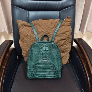 Puede incluir: Una mochila de color verde oscuro con un patrón texturizado en relieve que recuerda a la piel de reptil. La bolsa incluye un asa superior, dos correas para los hombros y un bolsillo frontal con cremallera y herrajes plateados. El texto Lucky Strike Handmade es visible.