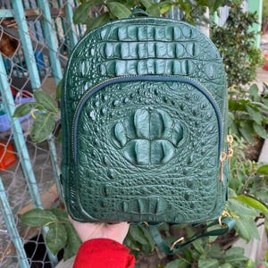 Op de afbeelding: Een donkergroene rugzak met een gestructureerd krokodillenleerpatroon. De tas heeft een afgeronde bovenkant, een voorvak met rits en een gebogen opening, en goudkleurige metalen ritstrekkers. Het oppervlak heeft een glanzende afwerking.