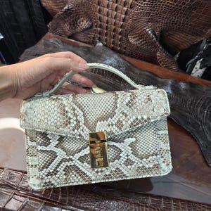 May include: A structured handbag crafted from snakeskin with a natural pattern of cream, beige, and brown scales. The bag features a top handle and a rectangular gold-tone metal combination lock closure on the front flap. The text Lucky Strike Handmade is visible.