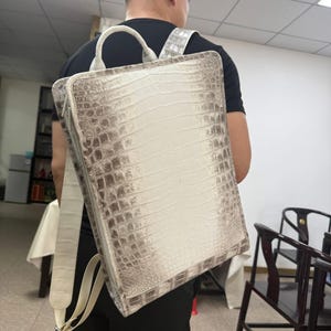 Op de afbeelding: Een vierkante rugzak met een gestructureerd krokodillenleerpatroon in gebroken wit en lichtbruin. De tas is voorzien van een handvat aan de bovenkant, verstelbare schouderbanden en een ritssluiting aan de zijkant. De tekst Lucky Strike Handmade is zichtbaar.