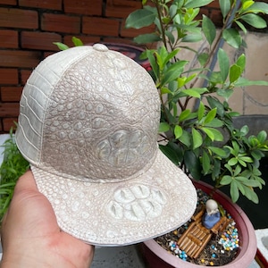 May include: A baseball cap crafted from textured off-white and beige leather with a distinct reptilian scale pattern. The cap features a structured crown and a flat brim, showcasing a prominent, large scale detail on both the front panel and the brim.