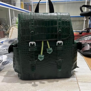 Op de afbeelding: Een donkergroene rugzak met een gestructureerd krokodillenleerpatroon. De tas is voorzien van een handvat aan de bovenkant, een overslagklep met twee zilverkleurige metalen gespen en twee lichtgroene trekkoorden met bijpassende lederen lipjes.
