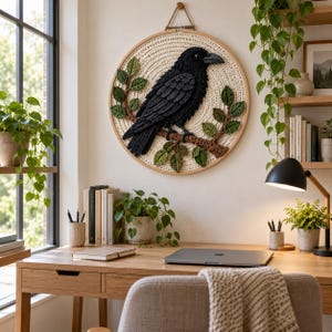 May include: A circular wall hanging featuring a crocheted black crow perched on a brown branch with green leaves. The crow is set against a cream-colored crocheted background within a wooden embroidery hoop, suspended by a string.