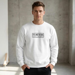 May include: A white long-sleeved crewneck sweatshirt featuring a rectangular graphic on the chest. The graphic contains the text "IT'S NOT A BUG IT'S A FEATURE" in a dark, distressed font, enclosed within a thin, rectangular border.