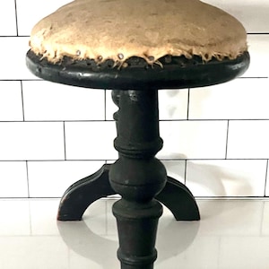 May include: A dark brown, antique stool with a round, padded seat. The seat is light tan and appears to be made of leather or a similar material. The stool has a turned central column and three curved legs.