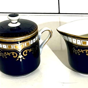 May include: A navy blue and gold tea set, featuring a covered sugar bowl and a creamer. The set has white interiors, gold trim, and decorative gold scrollwork. The sugar bowl has a white knob on the lid. The creamer has a curved spout.
