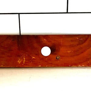 May include: A rectangular, wooden object with a warm, reddish-brown hue. It features a large, centered white circle and a smaller hole to the right. The wood grain is visible, and the surface shows signs of wear.
