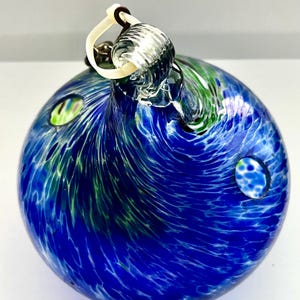 May include: A hand-blown glass ornament with a swirling blue and green design, resembling a miniature planet. The ornament has a clear glass loop for hanging and two small, round openings. The glass is textured with white streaks.