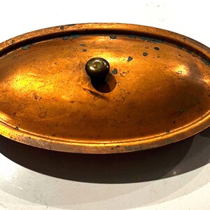 May include: Oval copper casserole dish with a lid and brass handles. The dish has a dark brown knob on the lid. The copper has a warm, aged patina. The handles are a bright gold color.