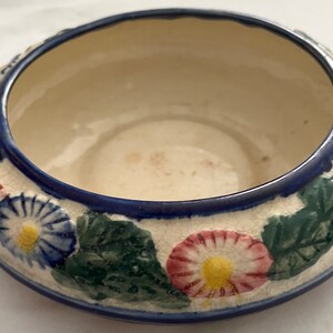 May include: A white ceramic bowl with a blue rim and floral design. The bowl features green leaves and pink, blue, and yellow flowers.