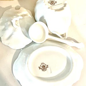 May include: A collection of white porcelain serving pieces, including a gravy boat, a ladle, and a plate. The pieces have a scalloped edge and a brown emblem that reads "Royal Semi-Porcelain England".
