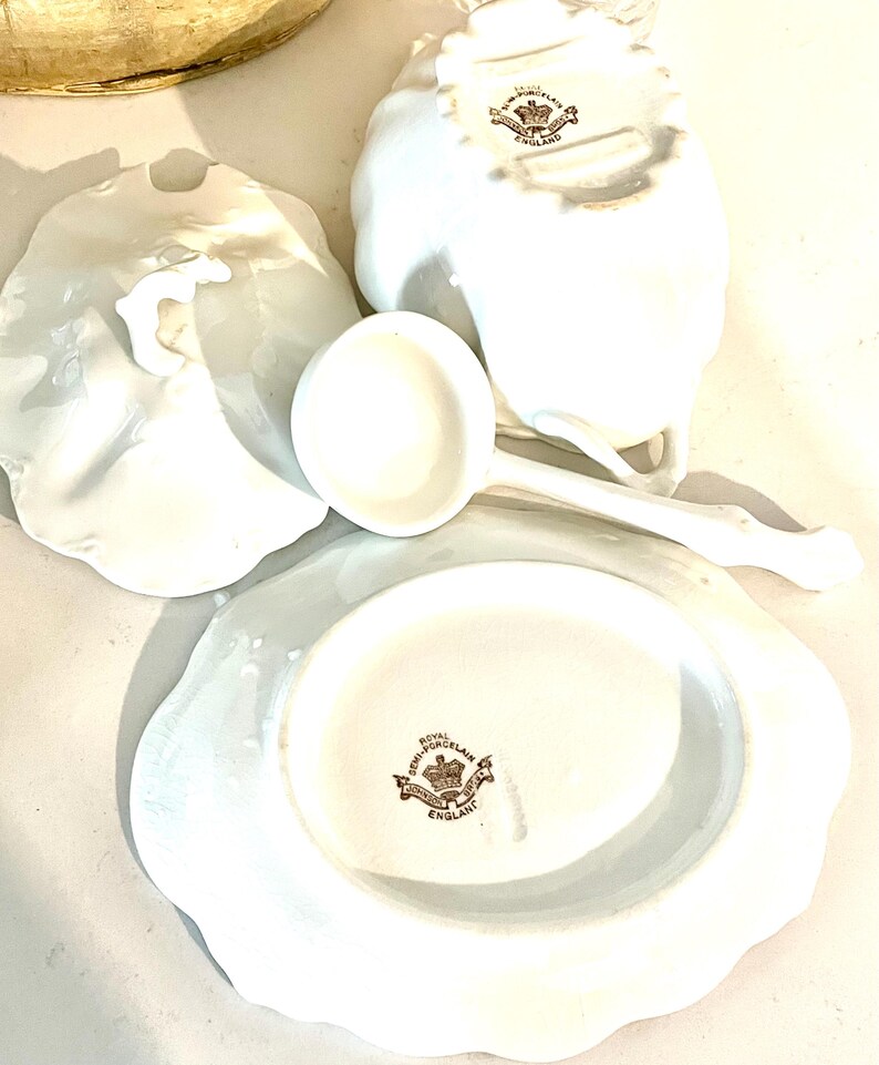 May include: A collection of white porcelain serving pieces, including a gravy boat, a ladle, and a plate. The pieces have a scalloped edge and a brown emblem that reads "Royal Semi-Porcelain England".