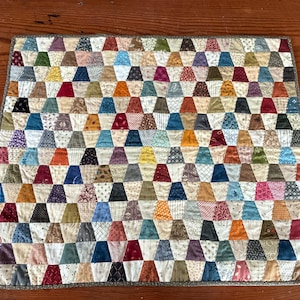 May include: A square fabric quilt featuring a repeating pattern of small, multi-colored trapezoidal patches. The quilt is composed of various printed cotton fabrics in shades of red, blue, yellow, green, brown, and cream, finished with a thin brown border.