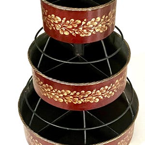 May include: A tiered organizer with three levels. Each level is a dark red with a gold floral design. The organizer has a black wire frame and a gold handle on top. The organizer is designed to hold small items.