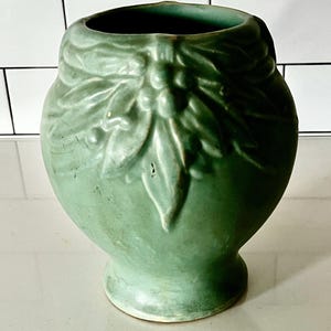 May include: A green ceramic vase with a matte finish. The vase features a detailed embossed floral design around the top and a rounded body. The vase has a small, circular base and a wide opening.