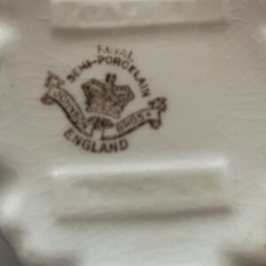May include: Close-up of a white porcelain dish with a scalloped edge. The bottom of the dish features a brown emblem with the words "Royal Semi-Porcelain" and "Johnson Bros England". The dish has a slightly aged appearance.