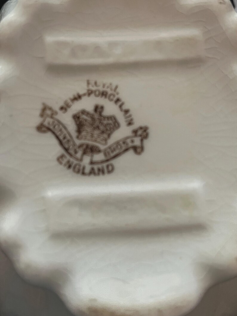 May include: Close-up of a white porcelain dish with a scalloped edge. The bottom of the dish features a brown emblem with the words "Royal Semi-Porcelain" and "Johnson Bros England". The dish has a slightly aged appearance.