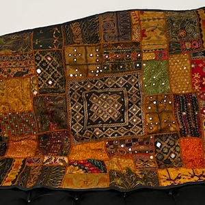 May include: A patchwork textile with a variety of colors including gold, orange, green, and brown. The textile is made up of many different squares and rectangles, with a variety of patterns and textures. The textile is draped over a black surface.