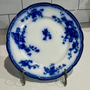 May include: White plate with a blue floral design and a scalloped edge. The plate features a central floral motif and additional floral patterns around the rim. The plate is displayed on a decorative metal stand.