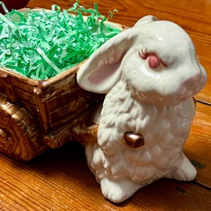 May include: A white ceramic bunny figurine pulling a brown wooden cart filled with green paper grass. The bunny has pink accents on its ears, nose, and closed eyes. A decorative Easter-themed item.